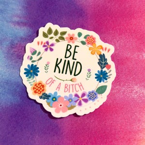 May include: A white sticker with a floral wreath design. The text "BE KIND OF A BITCH" is in the center. The sticker features various colorful flowers and green leaves, set against a vibrant pink and purple background.
