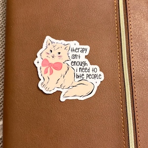 May include: A sticker featuring a cartoon cat with a pink bow, with the text "therapy isn't enough i need to bite people." The sticker is placed on a brown leather notebook with a zipper.