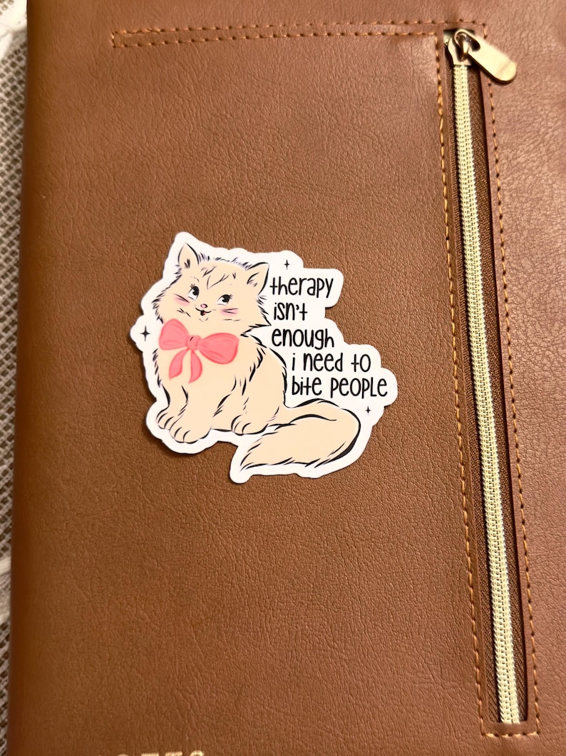 May include: A sticker featuring a cartoon cat with a pink bow, with the text "therapy isn't enough i need to bite people." The sticker is placed on a brown leather notebook with a zipper.