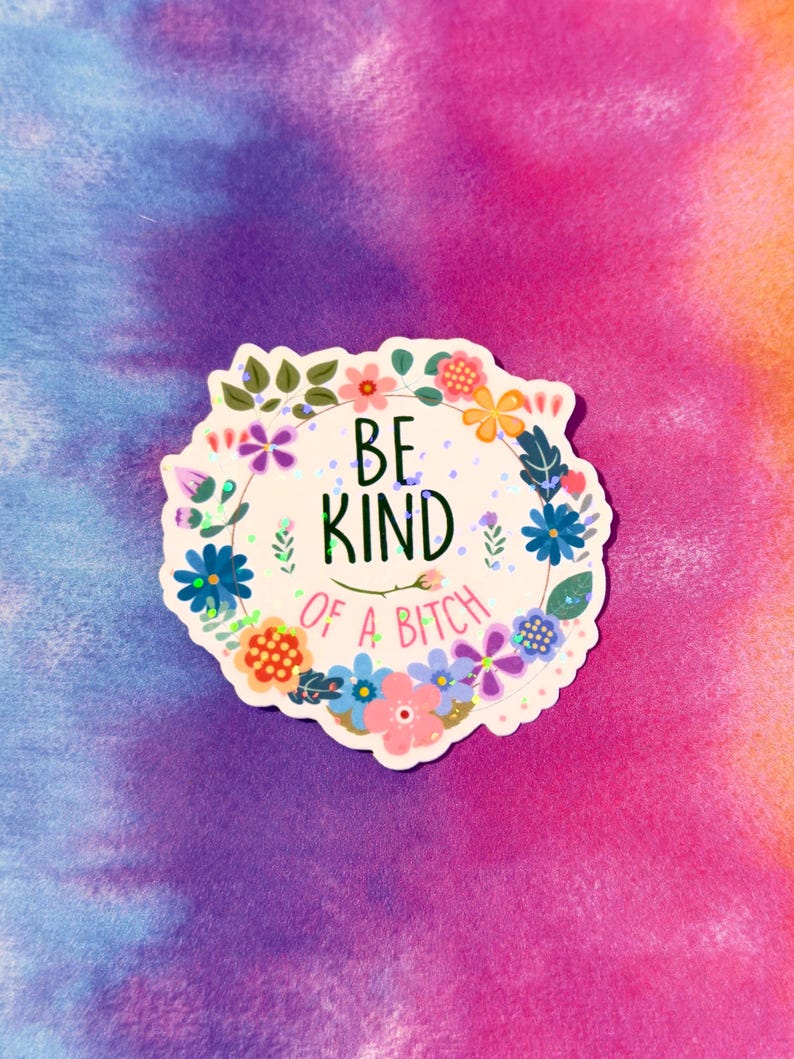 May include: A sticker with a floral wreath design and the text "BE KIND OF A BITCH". The sticker features a variety of colorful flowers and leaves, with a tie-dye background in shades of purple, pink, and orange.