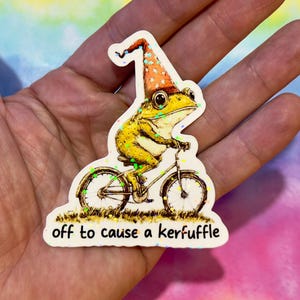 Könnte beinhalten: Ein skurriler Aufkleber mit einem gelben Frosch auf einem Fahrrad, der einen spitzen Hut mit orangefarbenen und weißen Punkten trägt. Der Text "off to cause a kerfuffle" ist darunter gedruckt.
