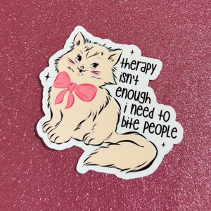 May include: A sticker featuring a cartoon cat with a pink bow and the text "therapy isn't enough i need to bite people." The cat is light tan with black outlines and is set against a pink glitter background.