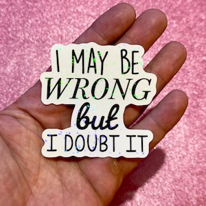 May include: A die-cut sticker with a white background and a holographic glitter finish. The sticker features the black text I MAY BE WRONG but I DOUBT IT in a combination of bold sans-serif and cursive script fonts, arranged in three distinct lines.