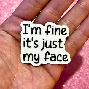 May include: A white sticker with the text "I'm fine it's just my face" in black, held in a hand. The sticker has a rounded, irregular shape. The background is a pink, glittery surface.