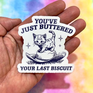 May include: A white sticker with a navy blue design featuring a kitten standing on a cowboy hat. The text "YOU'VE JUST BUTTERED YOUR LAST BISCUIT" is printed above and below the image of the kitten and hat.
