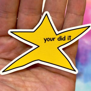 以下が含まれることがあります： 「your did it」の黒い文字が入った黄色の星型のステッカー。ステッカーは白い縁取りがあり、手で持たれています。背景はピンク、紫、青のカラフルなブレンドです。