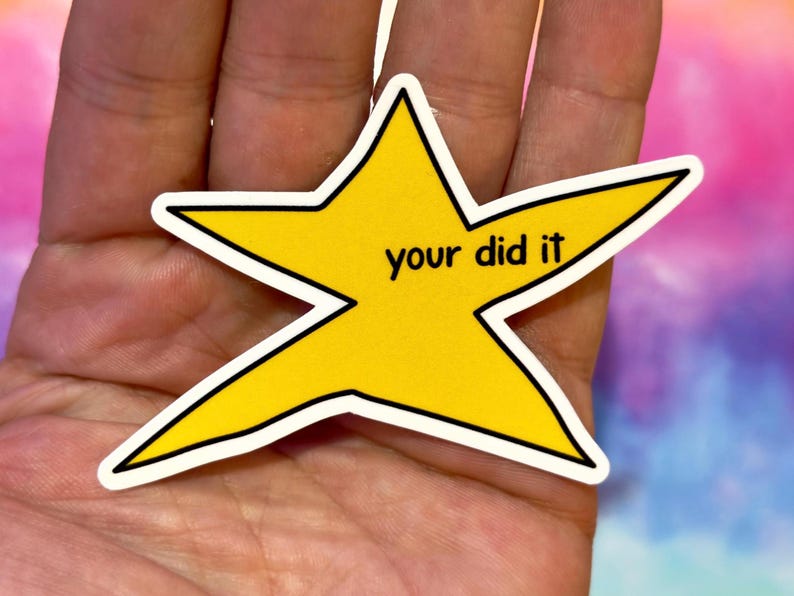 以下が含まれることがあります： 「your did it」の黒い文字が入った黄色の星型のステッカー。ステッカーは白い縁取りがあり、手で持たれています。背景はピンク、紫、青のカラフルなブレンドです。