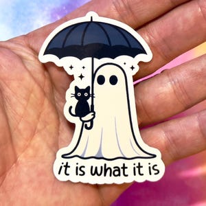 May include: A die-cut sticker featuring a cartoon ghost holding a black umbrella with a black cat. The ghost has two black eyes and is wearing a white sheet. The text "it is what it is" is at the bottom.