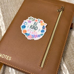 May include: A brown leather notebook with a gold zipper and the word "NOTES" embossed on the front. A sticker with the text "BE KIND of a BITCH" surrounded by a floral wreath is affixed to the cover.