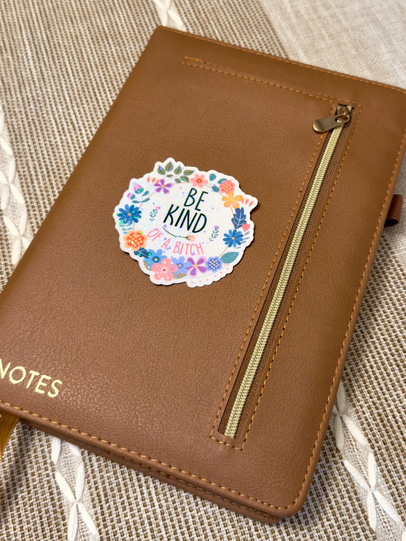 May include: A brown leather notebook with a gold zipper and the word "NOTES" embossed on the front. A sticker with the text "BE KIND of a BITCH" surrounded by a floral wreath is affixed to the cover.