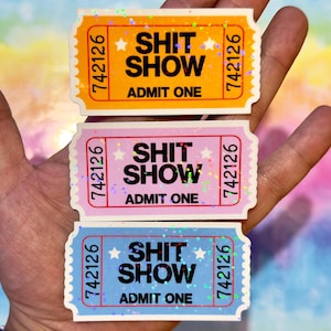 以下が含まれることがあります： 「SHIT SHOW ADMIT ONE」の文字が入った、チケット型のステッカー3枚。オレンジ、ピンク、青の3色で、それぞれ異なる配色と星のグラフィックが特徴です。背景は虹色です。