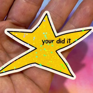 Op de afbeelding: Een gele ster-vormige sticker met de tekst "your did it" in zwarte letters. De ster heeft een zwarte omtrek en een witte rand. De ster is bedekt met glitter en heeft een kleurrijke achtergrond.