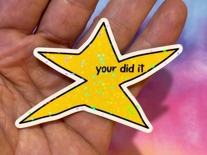 以下が含まれることがあります： 「your did it」の黒い文字が入った黄色の星型のステッカー。星は黒い輪郭と白い縁取りがあります。星はキラキラで覆われており、カラフルな背景があります。