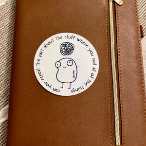 May include: A brown leather notebook with a gold zipper and the word "NOTES" embossed on the bottom left. A circular sticker with a cartoon figure and the text "Can you repeat the part about the stuff where you said all of the things" is on the cover.