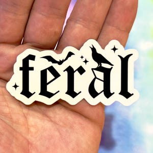 May include: A white sticker with the word "feral" in black gothic lettering. The sticker features a bat, a crow, and stars. The sticker is held in a hand.
