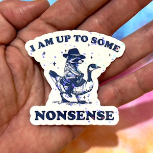 Op de afbeelding: Een sticker met een marineblauw ontwerp van een wasbeer met hoed en jas die op een gans rijdt, met de tekst "I AM UP TO SOME NONSENSE". De sticker heeft een holografisch glittereffect en een witte rand.