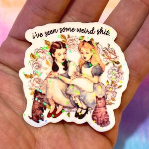 Könnte beinhalten: Ein weißer Aufkleber mit dem Text "i've seen some weird shit." Der Aufkleber zeigt eine Illustration von zwei Frauen in Kleidern, umgeben von Blumen und Katzen. Grüner Glitzer akzentuiert das Design.