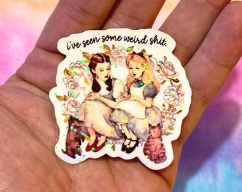 May include: A white sticker with the text "i've seen some weird shit." The sticker features an illustration of two women in dresses, surrounded by flowers and cats. Green glitter accents the design.