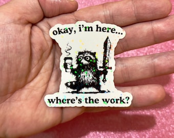 May include: A sticker featuring a cartoon raccoon holding a coffee cup and a sword. The raccoon is black and white with green accents. Text on the sticker reads "okay, I'm here... where's the work?"