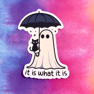 May include: A die-cut sticker featuring a cartoon ghost holding a black umbrella and a black cat. The text "it is what it is" is at the bottom. The background is a watercolor wash of purple, pink, and orange.