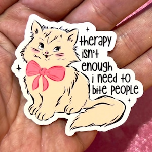May include: A sticker featuring a cartoon cat with a pink bow, with the text "therapy isn't enough I need to bite people." The cat is cream-colored with black outlines and whiskers. The sticker has a white border and black stars.