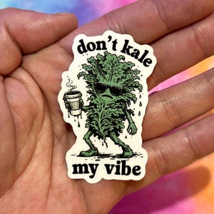 May include: A sticker featuring a cartoon kale character wearing sunglasses and holding a coffee cup. The text on the sticker reads "don't kale my vibe" in black font. The kale is green, and the sticker has a white border.