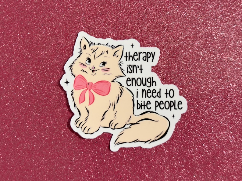 May include: A sticker featuring a cartoon cat with a pink bow, with the text "therapy isn't enough I need to bite people." The cat is beige with black outlines, set against a glittery red background.
