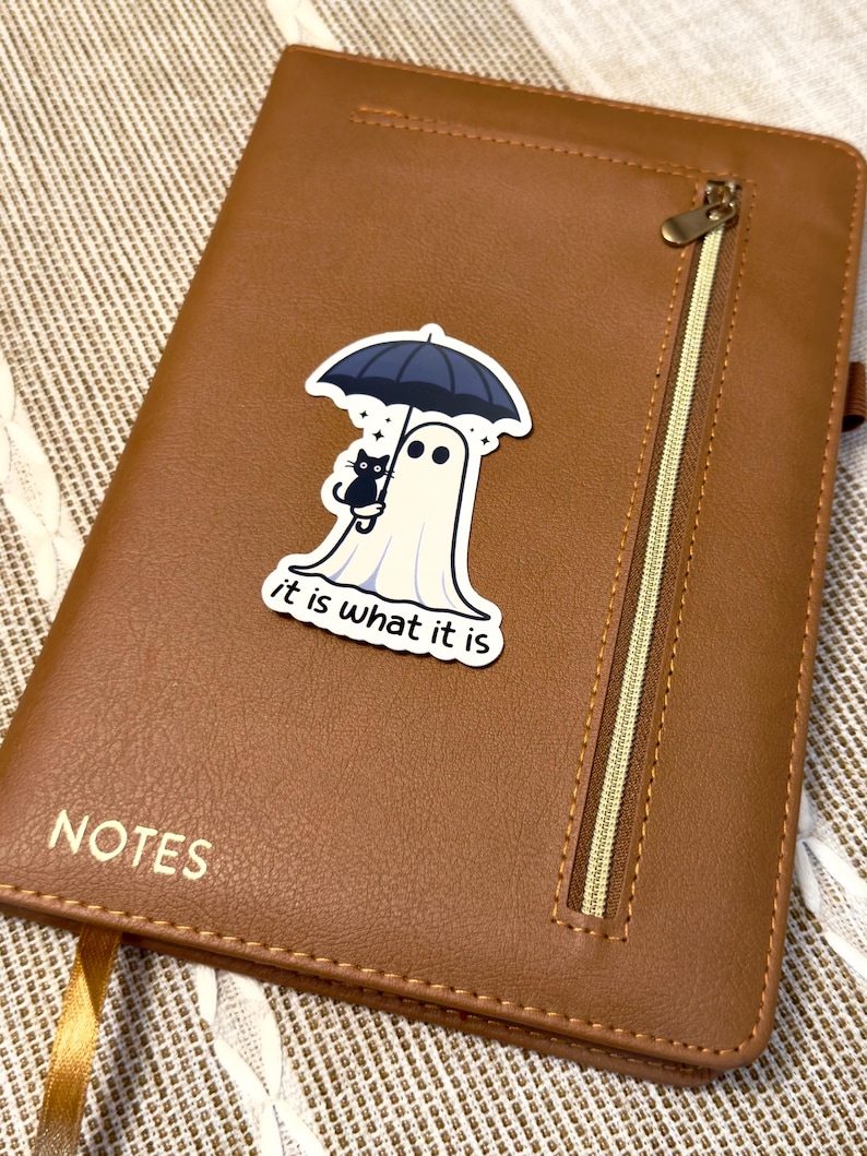 May include: A brown leather notebook with a zipper and the word "NOTES" embossed on the front. A sticker of a ghost holding an umbrella with a black cat is on the cover. The sticker has the text "it is what it is".