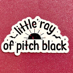 May include: A white sticker with the text "little ray of pitch black" in a semi-circle design. The sticker features a black sun with rays, bats, and stars. The background is a glittery, reddish-pink surface.