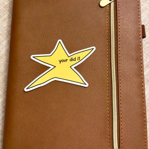 以下が含まれることがあります： 金色のジッパーと下部にエンボス加工された「NOTES」の文字が付いた茶色のフェイクレザーノート。 「your did it」という言葉が入った黄色の星のステッカーが前面にあります。 ノートにはステッチされたボーダーがあります。