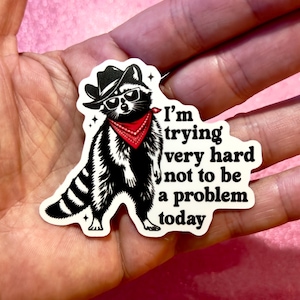 Puede incluir: Una pegatina de mapache en blanco y negro con sombrero de vaquero, gafas de sol y pañuelo rojo. La pegatina tiene el texto "I'm trying very hard not to be a problem today."