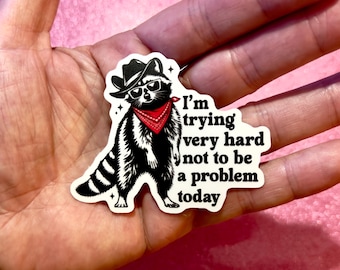 May include: A black and white raccoon sticker wearing a cowboy hat, sunglasses, and a red bandana. The sticker has the text "I'm trying very hard not to be a problem today."