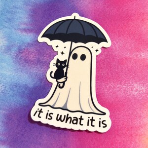 May include: A sticker featuring a cartoon ghost holding a black umbrella with a black cat. The ghost has two black eyes and is wearing a white sheet. The text "it is what it is" is at the bottom.