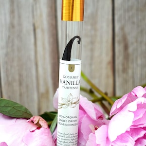 Puede incluir: Un tubo transparente con tapa dorada contiene una vaina de vainilla. La etiqueta dice "GOURMET VANILLA TAHITENSIS" y "100% ORGANIC SINGLE ORIGIN FROM INDONESIA". La vaina mide aproximadamente 19-20 cm. Flores rosas en primer plano.
