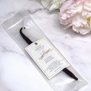 Puede incluir: Una vaina de vainilla única en un paquete de plástico transparente. La etiqueta dice "GOURMET VANILLA TAHITENSIS 100% ORGANIC SINGLE ORIGIN FROM INDONESIA". La vaina mide 19-20 cm. Flores blancas y rosas en el fondo.