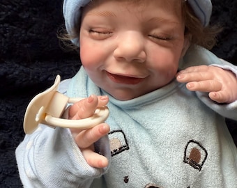 May include: A baby doll wearing a light blue beanie and matching onesie. The onesie features a bear face design. The doll is holding a pacifier and has closed eyes, with a slight smile. The doll's skin has a realistic tone.