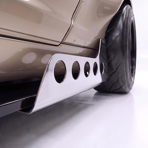 Puede incluir: Un panel de carrocería beige con un faldón lateral de cromo pulido con cinco recortes circulares, situado delante de un neumático negro con dibujo. El faldón lateral de metal está fijado al bastidor inferior con tornillos visibles.