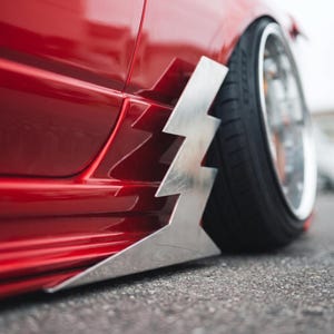 Peut inclure: Un panneau de carrosserie rouge vif doté d'une extension de bas de caisse métallique en forme d'éclair argenté, montée près de la roue avant. Le véhicule est équipé d'un pneu à profil bas avec une jante argentée polie sur une surface en asphalte.