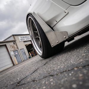 Puede incluir: Una aleta de coche blanca con un alerón lateral aerodinámico de metal plateado fijado con pernos visibles. La rueda presenta una llanta oscura de radios múltiples con un borde plateado pulido y un neumático negro. El coche está sobre asfalto.