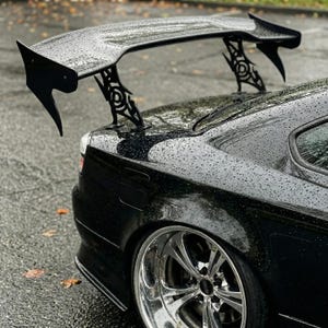 May include: Black car with a large, black spoiler. The spoiler is mounted on the boot and has decorative supports. The car has chrome rims and is wet from the rain.