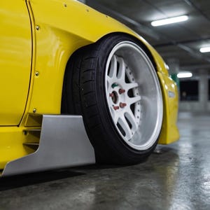 May include: A bright yellow car body panel featuring a wide fender flare with visible rivets, a metallic silver side skirt extension, and a large white multi-spoke wheel with a deep dish rim and black tire, captured from a low side angle.