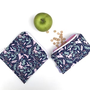 May include: Two reusable snack bags with a navy blue background and a pink and green floral pattern with unicorns. The bags have white zippers and are made of fabric.