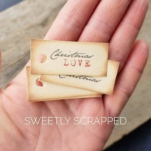 May include: A stack of three vintage-style gift tags with a distressed paper texture. The top tag features the text "Christmas LOVE" in black and red ink. The bottom two tags have the word "Christmas" written in black ink.