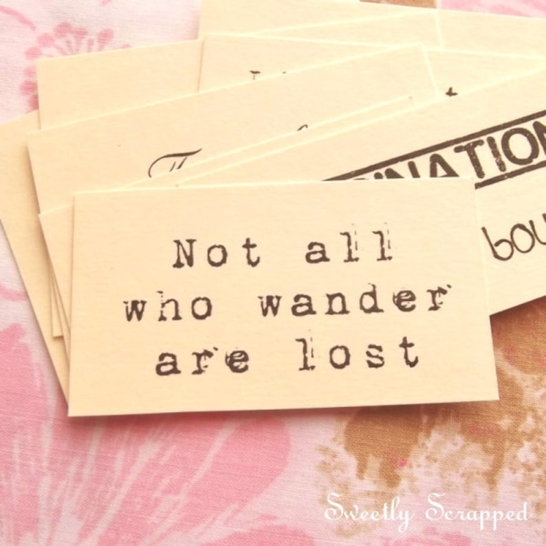 May include: A stack of cream-colored rectangular cards with black text. The primary card reads "Not all who wander are lost" in a typewriter font. Other cards have partial text. The background is a pink floral pattern.