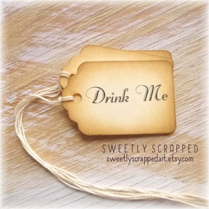 May include: Two vintage-style paper tags with scalloped edges and the words "Drink Me" printed in elegant script. The tags are tied together with a cream-colored string, set against a light wood background.