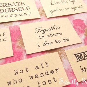 May include: Several small, cream-colored cards with black text are arranged on a floral background. The cards feature inspirational quotes like "Create Yourself Everyday" and "Together is where I love to be." The words are printed in a typewriter-style font.