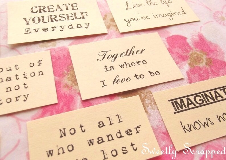 May include: Several small, cream-colored cards with black text are arranged on a floral background. The cards feature inspirational quotes like "Create Yourself Everyday" and "Together is where I love to be." The words are printed in a typewriter-style font.