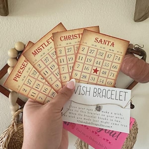 May include: A set of five vintage-style bingo cards with Christmas themes, including "Present", "Mistletoe", "Christmas", and "Santa". The cards are made of paper and have a cream background with red and black text. The cards are arranged in a fan shape and are held by a hand. A small card with the text "Wish Bracelet" is in the foreground. The card has a silver charm and the text "Close your eyes and make a wish, Then tie this bracelet on your wrist. When the bracelet breaks in two, The wish you made just might come true *DakotajadeDesigns.Etsy.com*"