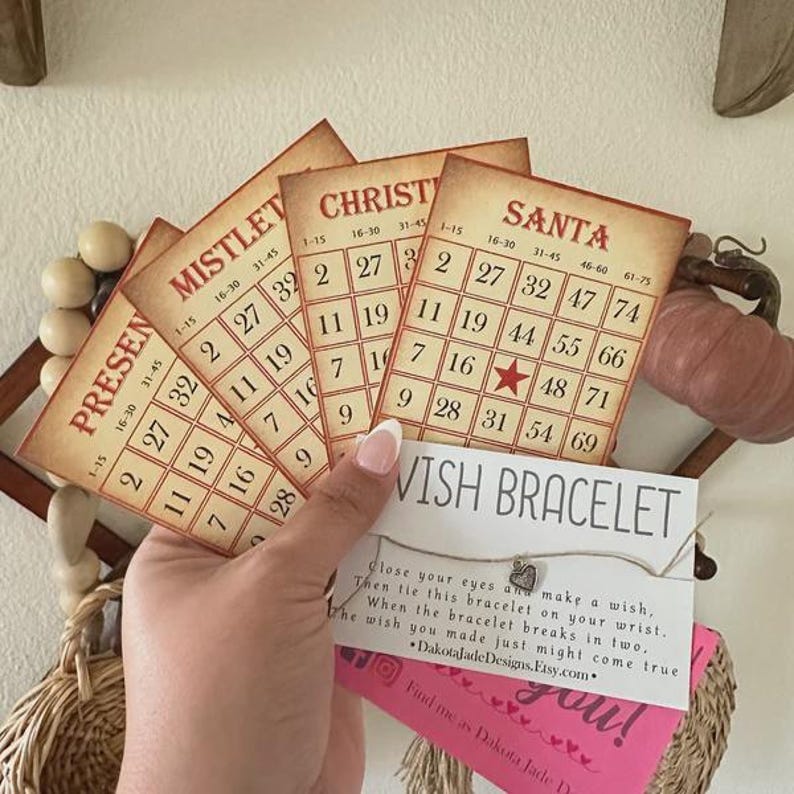 May include: A set of five vintage-style bingo cards with Christmas themes, including "Present", "Mistletoe", "Christmas", and "Santa". The cards are made of paper and have a cream background with red and black text. The cards are arranged in a fan shape and are held by a hand. A small card with the text "Wish Bracelet" is in the foreground. The card has a silver charm and the text "Close your eyes and make a wish, Then tie this bracelet on your wrist. When the bracelet breaks in two, The wish you made just might come true *DakotajadeDesigns.Etsy.com*"