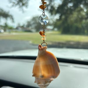 May include: A car charm featuring a natural seashell pendant. The charm is suspended by a chain with faceted crystal and amber-coloured beads. The seashell is a light orange colour.